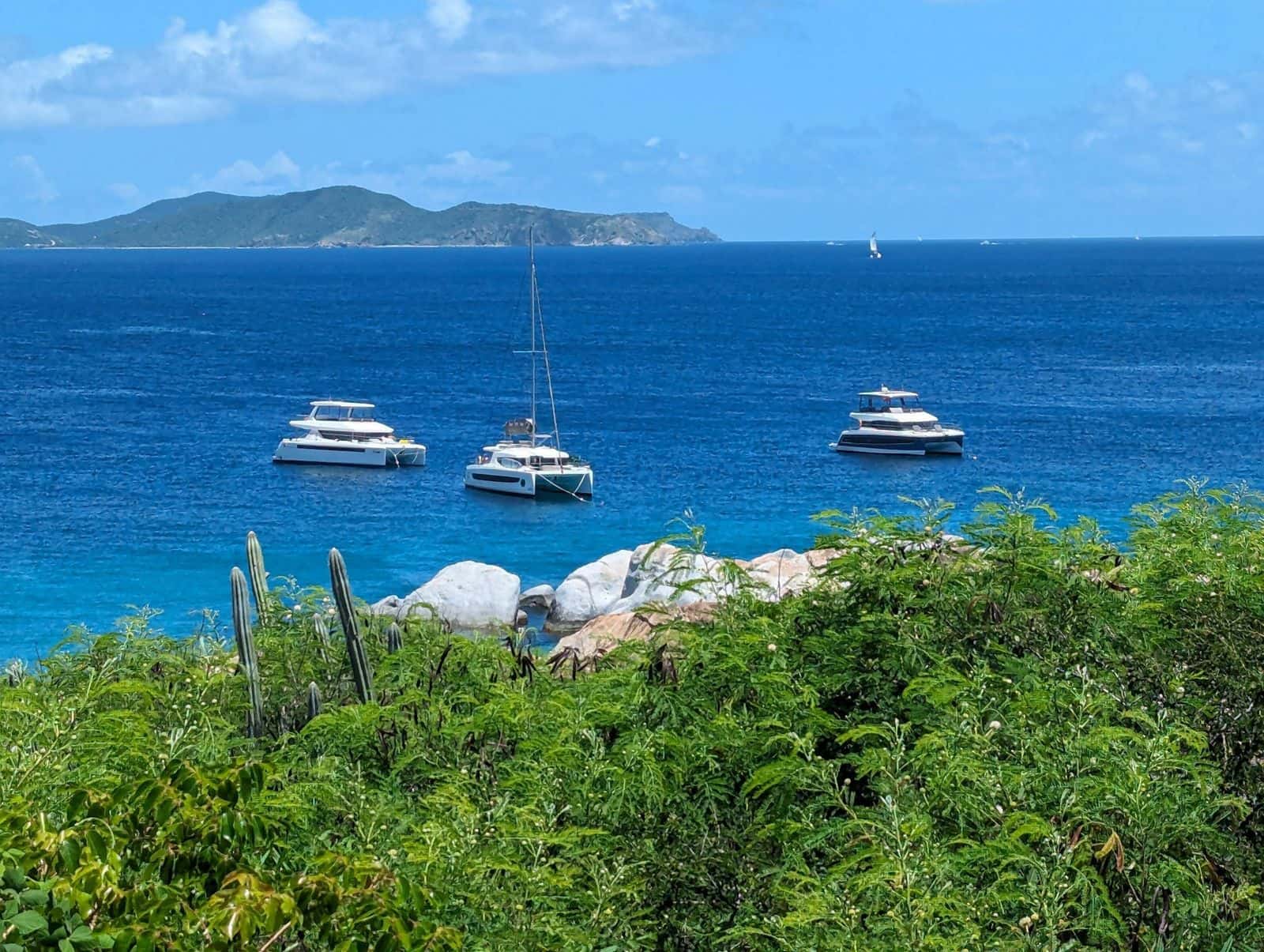 A sailboat and two catamarans anchored off a British Virgin Islands shore of boulders and cactus