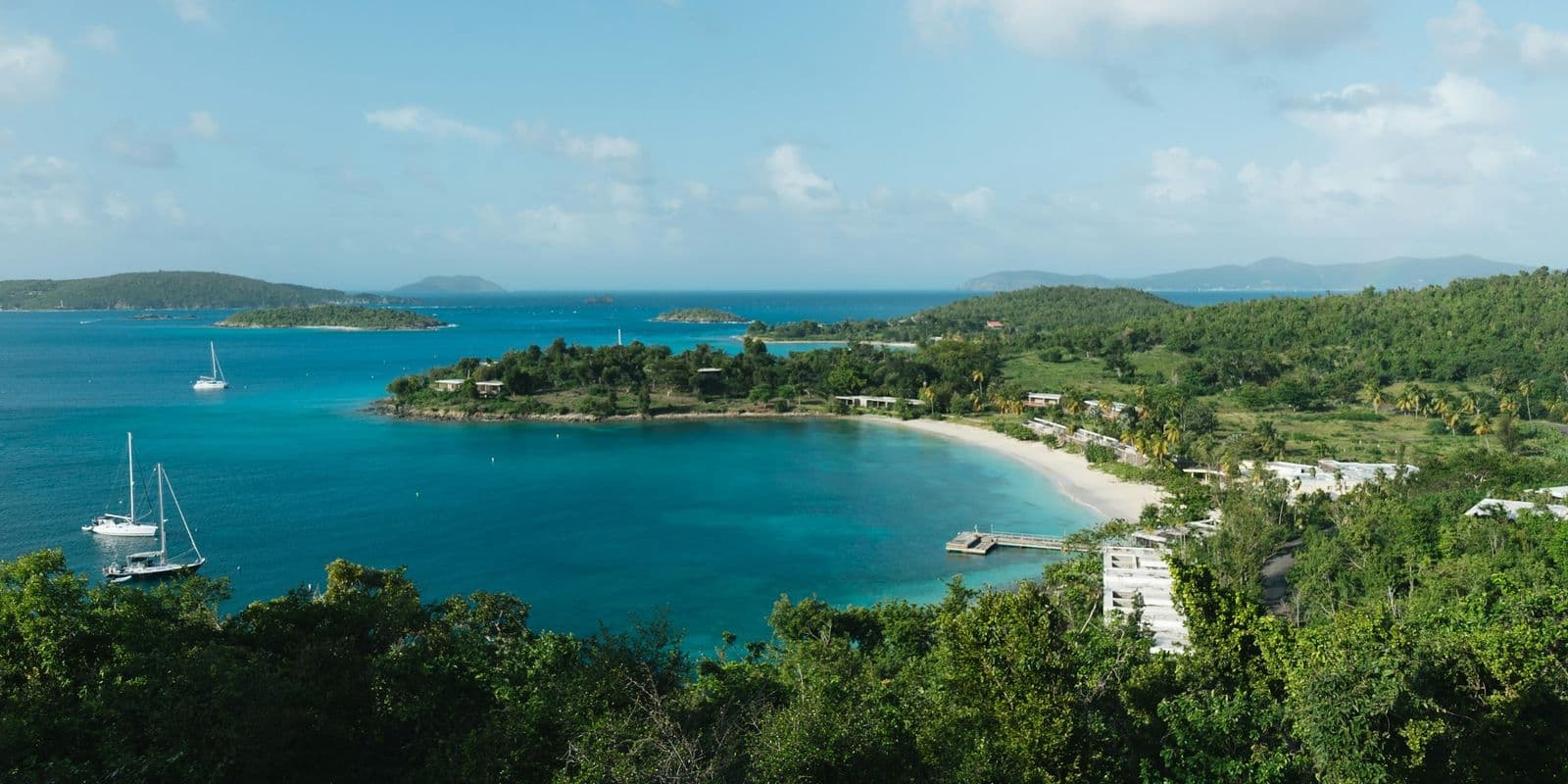A bay on St. John's north shore from the road overlook: a curved beach, a dock and low resort buildings, sailboats at anchor.