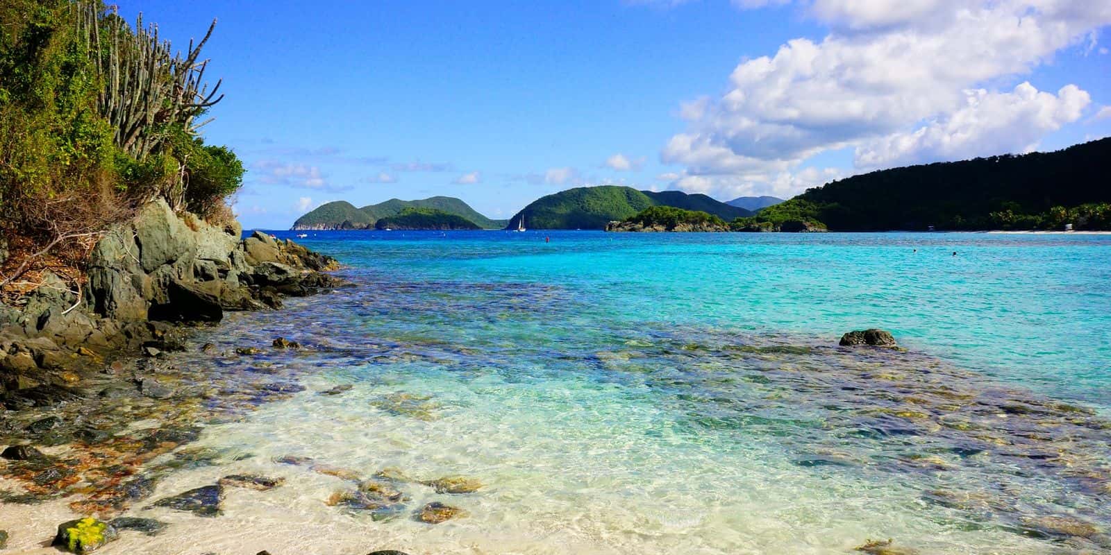 Clear shallows over rock at Jumbie Beach, St. John, seen from the sand, with a rocky point and cactus on the left.